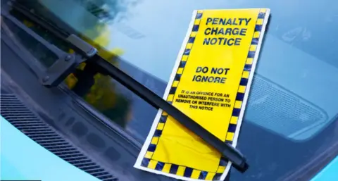 A yellow parking ticket which reads "do not ignore" slipped under a windscreen wiper of a car.