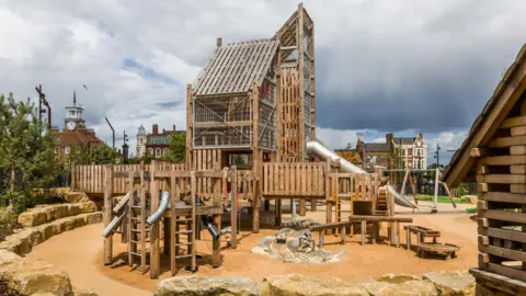 Stockton Council The wooden play area has two wooden towers and multiple slides and ladders coming off it. The structure is in a huge sand pit. There are swings in the background.