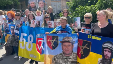 Families in northern Ukraine hold blue and yellow Ukrainian flags with pictures of their fathers and husbands