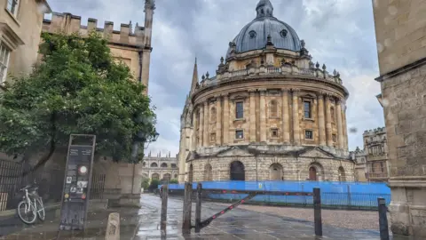 The Radcliffe Camera landmark in Oxford with rain covered pavements in front and a grey sky filled with clouds above