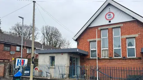 The image is of a number of buildings, including the Antrim branch of the Royal British Legion. It's pictured on the right and is a red brick building with tall windows and a Royal British Legion 100 emblem. It's surrounded by smaller red brick buildings and a blue metal fence. 