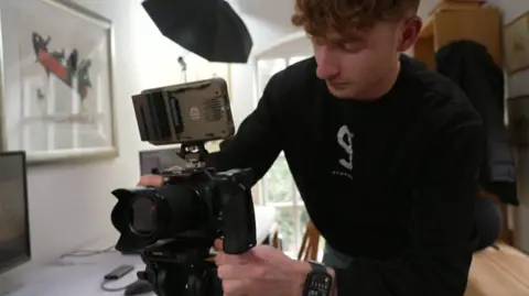A man wearing a black jumper stands usinga video camera. Other equipment and a desk can be seen in the background.