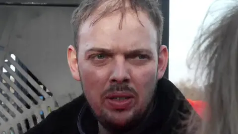 Chris Schofield with brown hair and beard wearing a black coat standing in front of machinery off an alleyway.