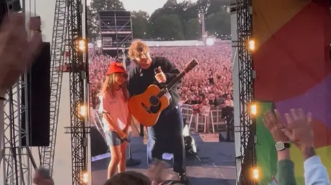 Evie-Mae stood next to Lewis Capaldi on stage. It is on the screen's stage and there are hands waving in the crowd.