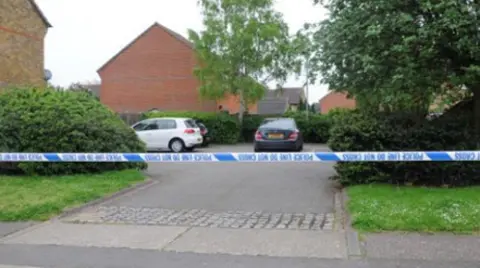 Essex Police A police cordon in Frobisher Way, Shoeburyness. Police tape is blocking off the entrance to a small residential car park, which has three cars parked inside it. The area is surrounded by trees and bushes.