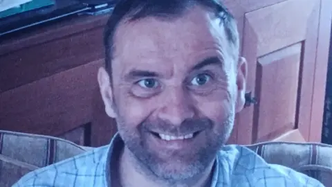 Police handout Mr Priestly is wearing a blue checked shirt and sitting on a striped settee with a wooden cabinet visible behind him. He is smiling at the camera.
