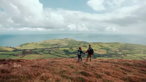 A man and a woman standing at the top of a green hill looking out towards the sea. They are wearing knitted jumpers, backpacks and hats, and are holding walking sticks.