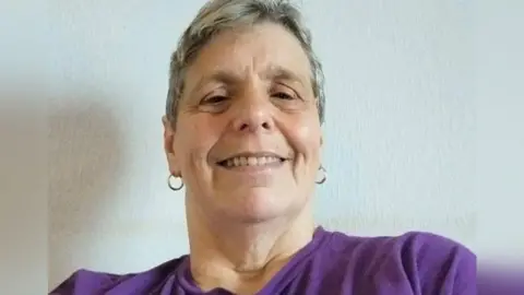 Family Handout A selfie of Sharon Hall. She has short grey hair and is wearing hoop earrings and a purple t-shirt.