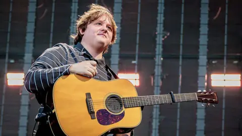 Getty Images Lewis Capaldi, who has long blonde hair and blue eyes, performs on stage with an acoustic guitar.