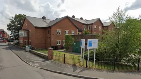 Google Exterior view of Sunnyview House care home. It is a brick building which looks like a house. There is a white sign outside with Bupa's blue logo on it.