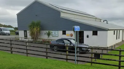 Google A picture of the community centre. It is a large white and grey building with cars parked around it and a large field next to it. 
