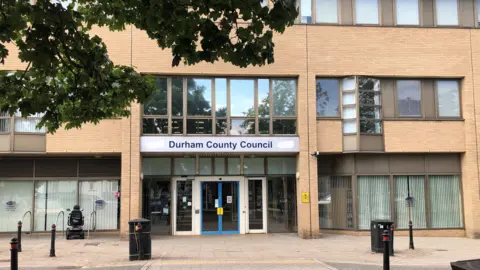 The front of an office building in light coloured brick facing onto a paved street. There are full windows with vertical blinds either side of a wide glass entrance door, with a sign for Durham County Council above it. Further windows are lined up on the first floor, and the part of the visible part of the second floor. Rubbish bins are either side of the entrance on the pavement, with the branches of a tree in full leaf on the top left.