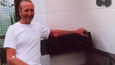 John Leigh John during his younger years. He is wearing a white t-shirt and is sitting on a bench smiling. 