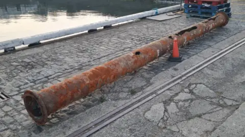 A rusted pipe with holes at the far end. It is lying on the floor at Guernsey's St Peter Port Harbour and the sea can be seen in the top left corner. 