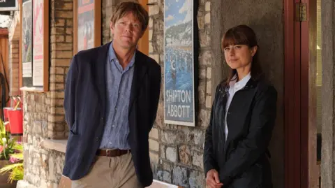 BBC/Joss Barratt A promotional photo of Beyond Paradise featuring actors Kris Marshall and Zahra Ahmadi. They are standing outside an old building with a sign saying "Shipton Abbot", the setting of the show. Kris Marshall has light brown hair and is wearing a dark navy blazer over a light blue button-up shirt and light beige trousers. He has hands behind his back. Zahra Ahmadi is standing in a doorway, leaning lightly against the wall. She has long dark hair and is wearing a dark, zip-up jacket over a white collared shirt. Her hands are in front of her.
