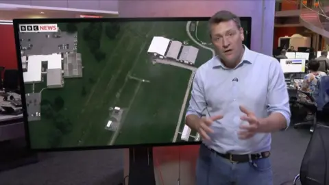 Joe Inwood stands in front of a TV screen showing an aerial of a green field with several buildings in it, the site of the attempted assassination 