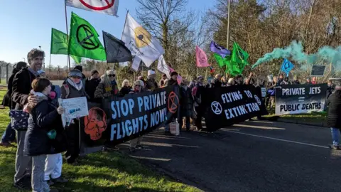 Campaigners with banners against the airport expansion standing outside the airport. The banners say stop private flights now, flying to extinction and private jets = public deaths.