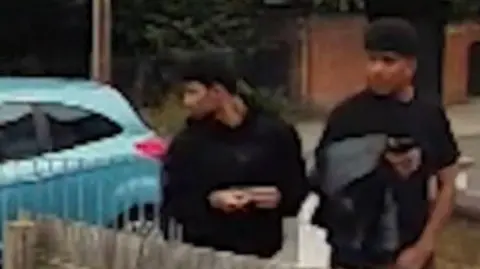 Two young men in black clothing are captured on CCTV looking to their right while standing in a residential area with a blue car behind them.
