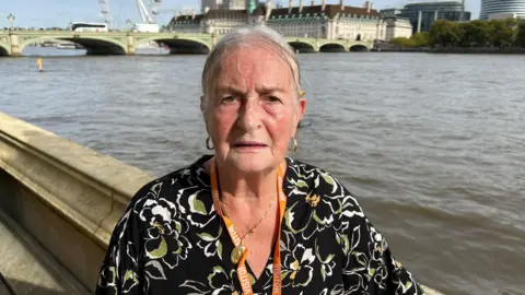 BBC/RICHARD EDWARDS Big Communi-Tea client Jackie Carroll is standing on the House of Commons terrace in front of the River Thames. The London Eye wheel is in the background. She is wearing a black blouse with gold and white flowers and a gold necklace.