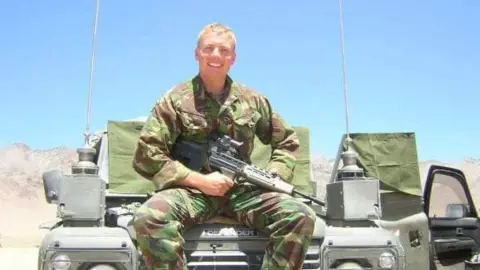 Christine Harrison Man in camouflage clothes with a gun, sits on a jeep in the desert. 