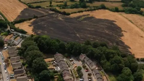 A blackened, scorched field in the countryside surrounded by brown and pale green fields behind and to the side. In front of the burnt field is a road lined by green trees and in front of that are residential roads and houses.