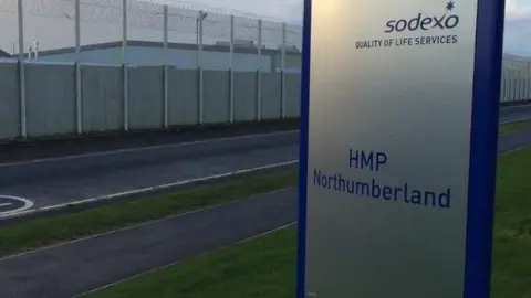BBC The outside of HMP Northumberland which is surrounded by a silver wire tall fence with barbed wire at the top. There is a road to the left and a large, silver sign in the foreground which reads 'HMP Northumberland' in blue letters. 