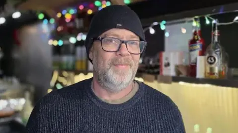Paul Moseley/BBC Hugo Malik, is standing inside his market stall, Churros for the People. He is wearing a black jumper and black beanie hat. He is looking directly at the camera and smiling.
