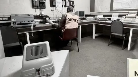 Nothe Fort Rear view of a man in a hi-vis vest sitting at a row of desks full of 1980s computer and telecoms equipment and machinery.