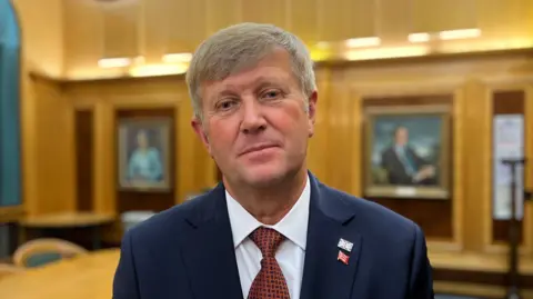 Peter Harris looking at the camera while standing in a wood-panelled council chamber. He is wearing a suit and tie. The tie is maroon and the blazer is navy blue. There are portraits on the wall in the background. He has two metal pin badges on a lapel - one of the union flag and one of the Essex triple seaxes.
