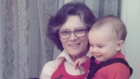 Peter Moone An old photo of a woman in glasses holding a baby boy in a red top. 