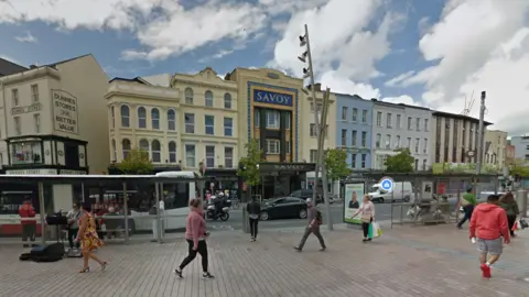 A Google Maps screen grab of St Patrick's St in Cork city centre.   There are shoppers walking along the street, parked cars and the Savoy cinema is in the background.