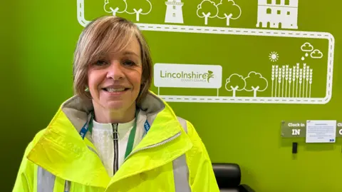 BBC News A woman wearing a high visibility jacket smiles at the camera. She is standing in front of a green wall with white illustrations and the words "Lincolnshire County Council".