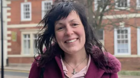 A person with dark, wavy hair stands on a quiet street lined with red‑brick buildings featuring white window frames. They are wearing a deep burgundy coat with a fur‑trimmed collar over a light pink top and a silver necklace. A tree with bare branches is visible behind them.
