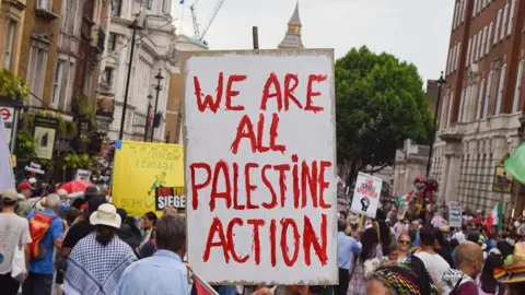 A protest in London - in focus is a sign which, in red paint, reads "We are all Palestine Action"