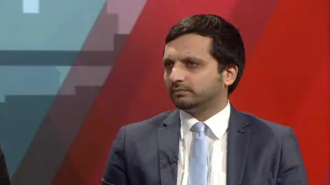 MP Saqib Bhatti  wears a grey suit, white shirt and light blue tie. He looks to the left of the camera with a solemn expression. He sits on a red sofa, with a blue backdrop behind him.