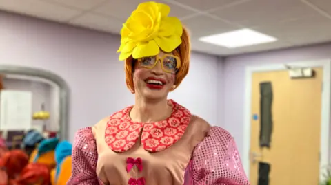 A man in a ginger wig, wearing make up, a large yellow flower and big glasses poses for the camera. He is in an ornate pink dress.
