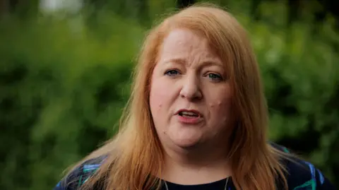 PA Media Headshot of Naomi Long looking at the camera a speaking with her eyebrows furrowed. Behind her some greenery that is out of focus.