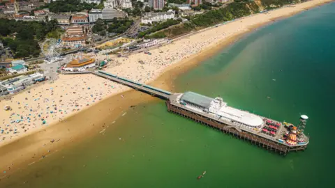 LDRS Aerial image of Bournemouth Pier with a large white building on it, a helter skelter and a high platform attached to a zip wire. The beach is busy and there is a big wheel on the pier approach.