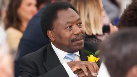 Professor Christopher Balogun-Lynch, sitting in a room, looking off to the right. He has a suit on, with a white shirt, blue tie, black jacket with a yellow rose on his lapel. He has short dark hair and has a moustache. 