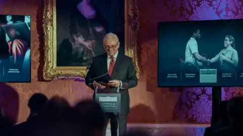 Amazon MGM Studios Michael Sheen, portraying Prince Andrew stands at a podium, smiling as he addresses a crowd of people, in front of posters and a portrait.