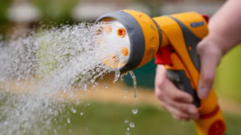A person holds a yellow and grey hosepipe with water spraying out of it. They are stood in a garden.