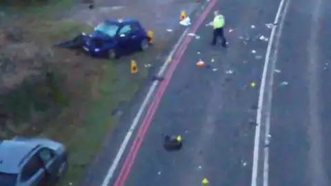 Avon and Somerset Police The scene of the crash with two cars off the road on the verge, one with a severely damaged bonnet. There are police cones around this car and a police officer in a high-vis jacket walking towards it. Other debris is scattered on the tarmac.