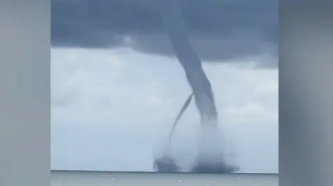 Two large dark grey waterspouts reach up into a dark cloud above the sea off the Italian coastline.