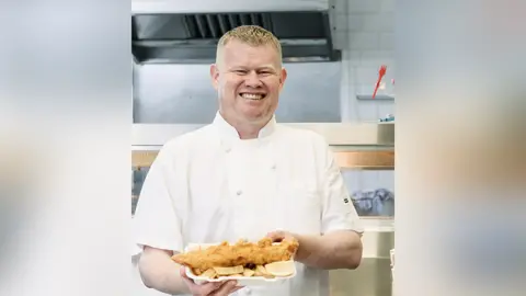 Andrew Crook handout Andrew Crook with blond hair wearing chef whites holding a portion of fish and chips with a wedge of lemon in a kitchen. He is smiling.