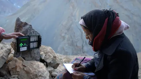 Devesh Chopra/BBC A woman wearing a black and red scarf writes something in her notebook and a camera trap is placed in front of her. 