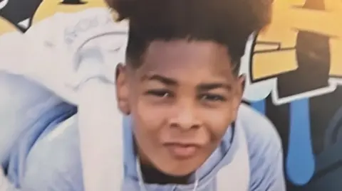 A photo of Kelyan, a boy of around 13, facing the camera. He has a high top haircut, and is wearing a light blue hoodie and white backpack. 