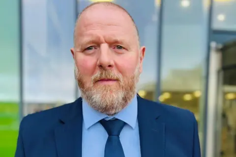 Man with beard and wearing light blue shirt, dark blue tie and dark blue suit jacket, looking at camera, with large glass windows in background.