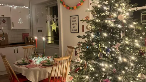 IWC Media Cozy kitchen and dining area decorated for Christmas with a large lit tree adorned with an eclectic mix of baubles, festive table setting, and a colourful paper garland on the wall.