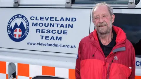 An older man stands next to a mountain rescue vehicle. He is wearing a red waterproof jacket with a small mountain logo on the chest, a black collared top