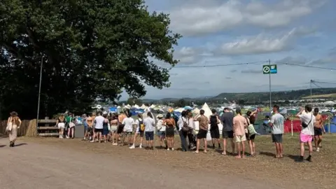 A queue of people waiting for their turn to fill up their water supply. There are tents in the background.